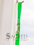 A fluro green biothane waterproof leash with brass hardware hanging from a brass hook on a white painted wooden post. It shows the top half of the leash with the leash clip attached to the D ring which is at the leash handle. The sky is in the background and there is a grey Sacred Pet Boutique watermark the bottom. 