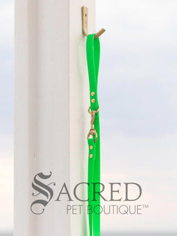 A fluro green biothane waterproof leash with brass hardware hanging from a brass hook on a white painted wooden post. It shows the full length of the leash which has it folded in half and the leash clip attached to the D ring at the leash handle. There is a beach in the background and a white Sacred Pet Boutique watermark the bottom. 