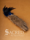 A single Da Fur Thing cat toy with brown fur and blue feathers coming out the top, lying on a wooden background with a white Sacred Pet Boutique watermark at the bottom of the image. 