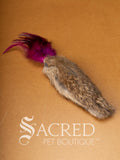 A single Da Fur Thing cat toy with brown fur and pink feathers coming out the top, lying on a wooden background with a white Sacred Pet Boutique watermark at the bottom of the image. 