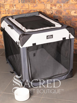 A Canine Care portable soft crate sits on a tile floor in a brick walled room. The side and an end door have been rolled up. An Italian Greyhound (it's Rossi) sits inside and looks to a person off screen. The crate looks spacious and airy.