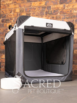 A Canine Care portable soft crate sits on a tile floor in a brick walled room. The side and an end door have been rolled up. An Italian Greyhound (it's Rossi) sits inside and looks to a person off screen. The crate looks spacious and airy.