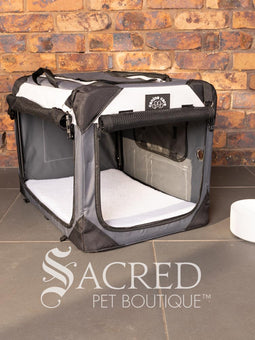 A Canine Care portable soft crate sits on a tile floor in a brick walled room. The side and an end door have been rolled up. An Italian Greyhound (it's Rossi) sits inside and looks to a person off screen. The crate looks spacious and airy.