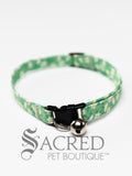A cat collar is shown against a plain white background. The pattern of the fabric is green, with darker green leaves and white daisy flowers. The collar has a black plastic breakaway buckle and adjuster, and a silver bell on a silver split ring.