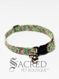 A cat collar is shown against a plain white background. The pattern of the fabric is green, with darker green leaves and a variety of dark and pale pink flowers with occasional small yellow flowers. The collar has a black plastic breakaway buckle and adjuster, and a silver bell on a silver split ring.