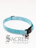 A cat collar is shown against a plain white background. The pattern of the fabric is mid blue with a crack pattern in paler blue. The collar has a black plastic breakaway buckle and adjuster, and a silver bell on a silver split ring.