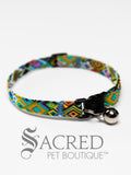 A cat collar is shown against a plain white background. The pattern of the fabric has many different sized and coloured diamond or lozenge shapes. The collar has a black plastic breakaway buckle and adjuster, and a silver bell on a silver split ring.