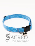 A cat collar is shown against a plain white background. The pattern of the fabric is mid blue, with a mix of darker and paler blue leaves and buds. The collar has a black plastic breakaway buckle and adjuster, and a silver bell on a silver split ring.