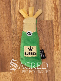 Three bottle shaped plush cat toys resting on a rustic wooden background. From the left there is a Rose, then a Whiskey and then Bubbly bottle. All three toys feature a fringed fabric top, a bottle shaped body and a black Zippy Claws label on the side. There is a white watermark of Sacred Pet Boutique at the bottom of the image. 