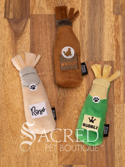 Three bottle shaped plush cat toys resting on a rustic wooden background. From the left there is a Rose, then a Whiskey and then Bubbly bottle. All three toys feature a fringed fabric top, a bottle shaped body and a black Zippy Claws label on the side. There is a white watermark of Sacred Pet Boutique at the bottom of the image. 