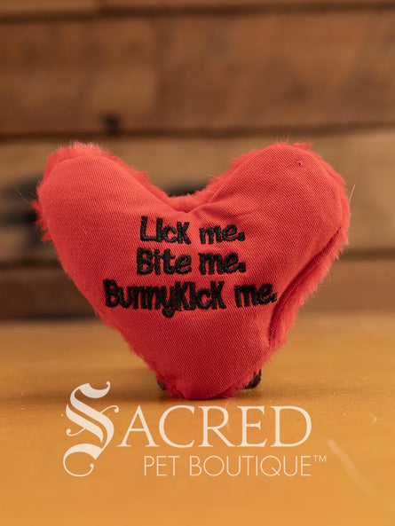 A red heart-shaped red catnip toy with text 'Lick me. Bite me. BunnyKick me.' embroidered on it in black. It is has faux fur on one side and plain material on the other. It is placed on a wooden surface with a white Sacred Pet Boutique watermark. 