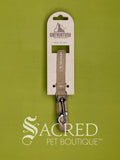 A tan pet leash with a silver metal clasp, attached to a kraft cardboard hanger displaying the Catventure brand. The product is set against a solid olive green background with a 'Sacred Pet Boutique' watermark at the bottom.