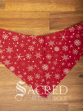 Red dog bandana with white snowflake pattern on a wooden surface, featuring the 'Sacred Pet Boutique' logo.