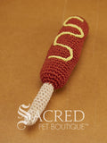 A cute crocheted hot dog on a stick with mustard lies on a wooden table with a wooden wall behind. Suitable for small, toy and teacup dogs, the crochet toys squeak and will floss teeth as they play.
