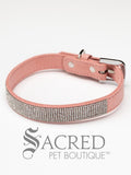 A pink faux suede dog collar with sparkling rhinestone accents and a rhinestone-encrusted buckle on white background and Sacred Pet Boutique watermark.