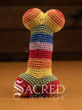 A cute crocheted rainbow coloured bone stands on a wooden table with a wooden wall behind. Suitable for small, toy and teacup dogs, the crochet toys squeak and will floss teeth as they play.