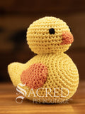 A cute crocheted yellow duck, like a rubber duck, sits on a wooden table with a wooden wall behind. Suitable for small, toy and teacup dogs, the crochet toys squeak and will floss teeth as they play.