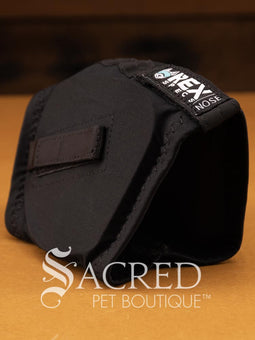 A black, hood-like sleeve made from a flexible 4-way stretch fabric that conforms snugly to a dog's head. This specialised hearing protection sleeve is designed to shield dogs from loud noises such as thunder, fireworks and gunfire. On the sides are small rectangular nylon loops to helps with stable adjustments or integrating with other tactical dog gear. The product is sitting on a wooden base and there is a white Sacred Pet Boutique watermark. 
