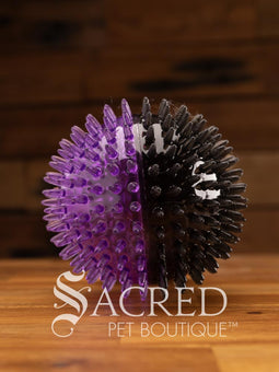 All three sizes of the squeaky spiky Galaxy ball are lined up for comparison. The balls are in two colour combinations - pink with orange, blue with green, and purple with black. All sitting on a wooden table with a wood plank background wall.