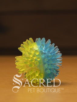 All three sizes of the squeaky spiky Galaxy ball are lined up for comparison. The balls are in two colour combinations - pink with orange, blue with green, and purple with black. All sitting on a wooden table with a wood plank background wall.