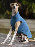 An Italian Greyhound dog sitting on a wooden platform in an out of focus outdoor setting wearing a Marine blue Goldpaw Stretch Fleece. A Sacred Pet Boutique watermark across the bottom.