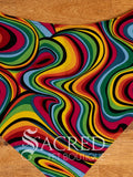 A large retro rainbow swirl dog bandana spread flat on wooden table and with a white Sacred Pet Boutique watermark