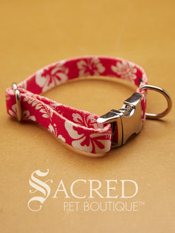 A red and white floral patterned webbed material pet collar with a silver metal buckle and D-ring, displayed on a warm tan background with the Sacred Pet Boutique watermark at the bottom.
