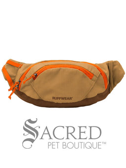 A person wearing a backpack with a blue and teal colour scheme is holding a treat in front of a dog's face. The dog is wearing a teal green harness and is attentive to the treat.