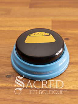 The product pack for Hunger for Words Talking Pet Essential Words sits on a wooden table with a wooden plank background. It shows the six pre-recorded blue and black buttons included, the words that are recorded, and pictures of dogs, although this product is suitable for cats.