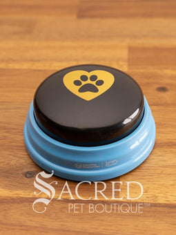 The product pack for Hunger for Words Talking Pet Essential Words sits on a wooden table with a wooden plank background. It shows the six pre-recorded blue and black buttons included, the words that are recorded, and pictures of dogs, although this product is suitable for cats.