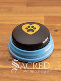 The Hunger for Words Talking Pet Essential Words "Love You" Button sits on a wooden table with a wooden plank background. The image of a bowl with a heart and paw print in it is clearly shown.