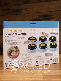 The product pack for Hunger for Words Talking Pet Essential Words sits on a wooden table with a wooden plank background. It shows the six pre-recorded blue and black buttons included, the words that are recorded, and pictures of dogs, although this product is suitable for cats.