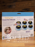 The back of the product pack for Hunger for Words Talking Pet Essential Words sits on a wooden table with a wooden plank background. It shows the six pre-recorded blue and black buttons included and the words that are recorded.