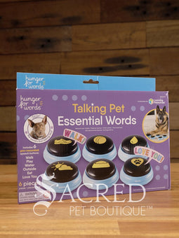 The product pack for Hunger for Words Talking Pet Essential Words sits on a wooden table with a wooden plank background. It shows the six pre-recorded blue and black buttons included, the words that are recorded, and pictures of dogs, although this product is suitable for cats.