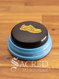 The Hunger for Words Talking Pet Essential Words "Walk" Button sits on a wooden table with a wooden plank background. The image of a shoe is clearly shown.