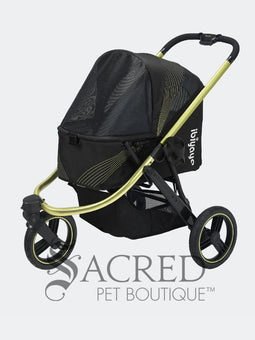 A woman in black athletic wear pushes a black pet stroller with green frame and wheel trim, in an outdoor urban setting. The stroller canopy has the brand name 'Ibiyaya' visible on the side. A white watermark from "Sacred Pet Boutique".