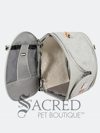 Person carrying a pet carrier with a cat inside, with a dog. surrounded by greenery, featuring the watermark 'Sacred Pet Boutique'.