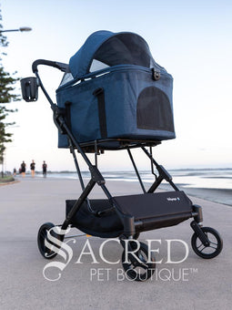 Pet stroller with a blue pet carrier on a paved path near a beach, featuring the Sacred Pet Boutique logo.