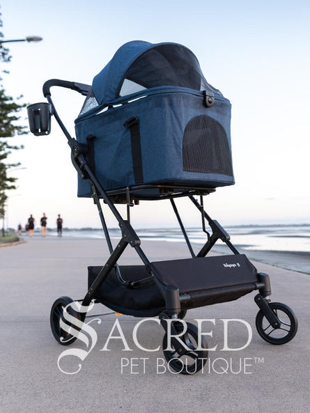 Pet stroller with a blue pet carrier on a paved path near a beach, featuring the Sacred Pet Boutique logo.