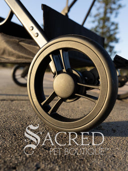 Pet stroller with a blue pet carrier on a paved path near a beach, featuring the Sacred Pet Boutique logo.