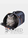 A gray long-haired cat sits inside a dark navy pet carrier bag with orange accents, shown against a light grey background. With the Sacred Pet Boutique logo at the bottom.