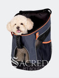 A small fluffy cream coloured dog sits inside a dark navy blue pet carrier backpack with orange trim, shown against a light grey background. A 'Sacred Pet Boutique' watermark is visible at the bottom.