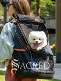 A woman outdoors wearing a navy blue pet backpack carrier with orange trim, with a small white dog peeking out from the top. The image has a 'Sacred Pet Boutique' watermark overlay.
