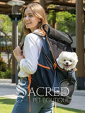 A woman outdoors wearing a navy blue pet backpack with orange accents, with a small white dog visible in the front pocket. The Sacred Pet Boutique logo is overlaid at the bottom.