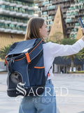A woman taking a selfie outdoors wearing a navy blue pet backpack with orange accents and a small dog visible in the front mesh pocket. The background shows a modern outdoor plaza with buildings and palm trees. With a white "Sacred Pet Boutqiue" watermark at the bottom.