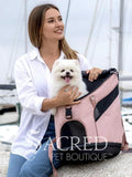 A woman in a white shirt and jeans holds a small white dog in a pink and black pet carrier backpack, set against a blurred marina background. The brand name 'Sacred Pet Boutique' is watermarked across the bottom.