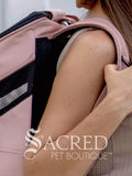 A close-up lifestyle shot of a person wearing a pink pet carrier backpack with black mesh panels, with the watermark'SACRED PET BOUTIQUE' overlaid at the bottom.