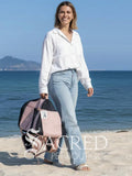 A woman standing on a sandy beach holds a pink and black pet carrier bag. She is wearing a white button-down shirt and light blue jeans, with the blue ocean and distant mountains in the background. A white Sacred Pet Boutique watermark at the bottom.