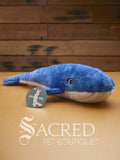 A dark blue and white whale sits on a wooden table with a wood panel wall in the background. It has embroidered eyes and furrows on its belly.