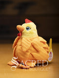 A bright yellow chicken with overlapping fabric feathers, flappy feet and embroidered eyes sits on a wooden surface. A white Sacred Pet Boutique watermark.